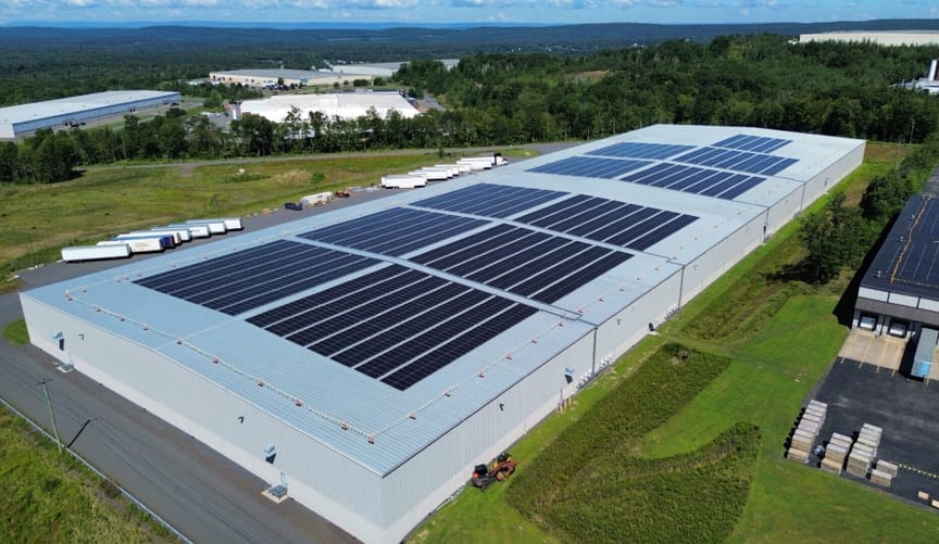 A large gray building, perhaps of corrugated steel, with solar panels on its gently sloped roof, on a grass lot