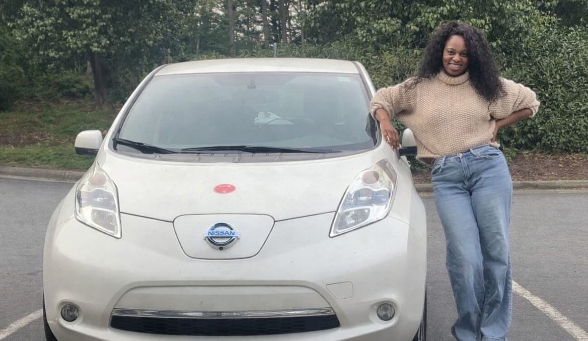 Woman in tan turtleneck and jeans leans against her 2013 Nissan Leaf.