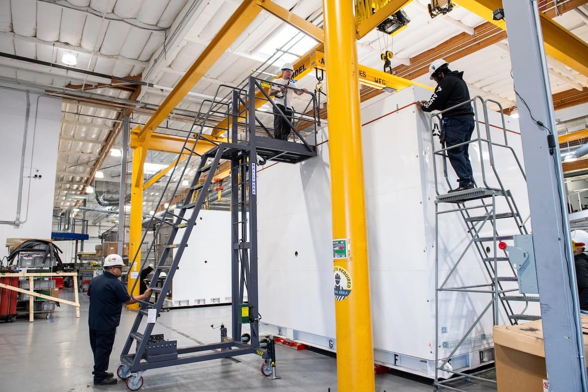 Two workers in white hard hats on ladders by white battery container