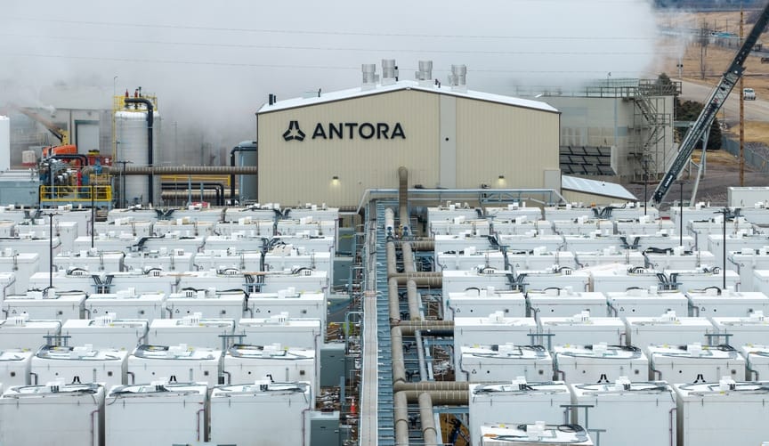 Rows of white batteries in front of a light beige building with "Antora" and its logo; steam rising in background