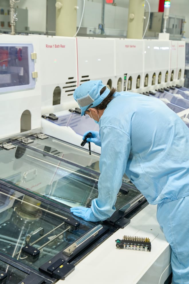 Blue-scrub-suted worker inspects a glass panel with a flashlight.