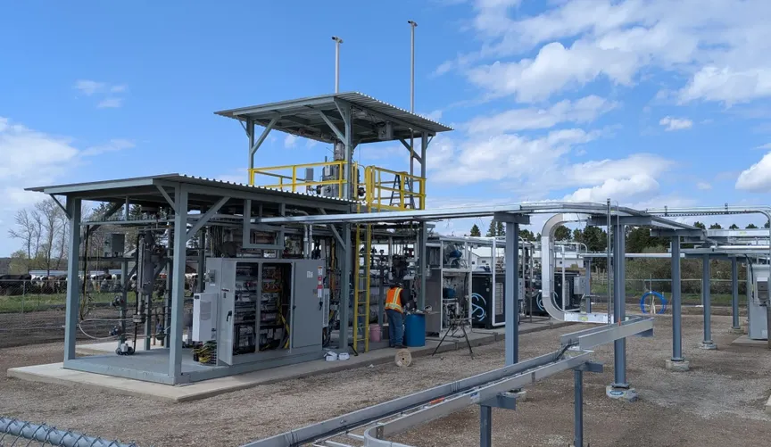Gray tanks, pipes, and sheds on a dirt plot; one orange-vested worker in the center