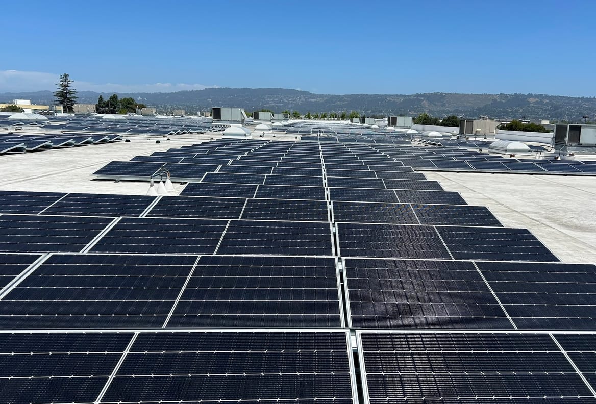 A large rooftop with many solar panels; hills and a blue sky in the distance