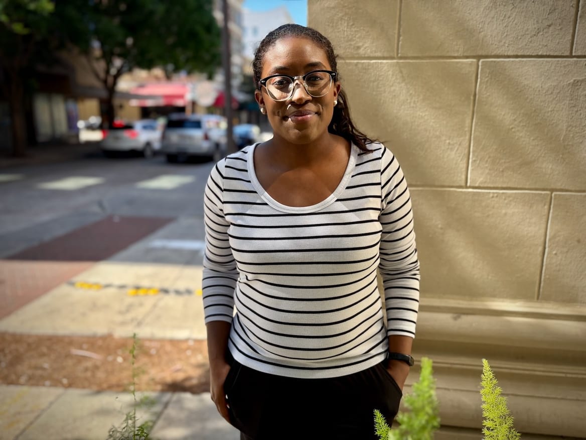 Woman in a white shirt with dark stripes stands at the corner of a building outside