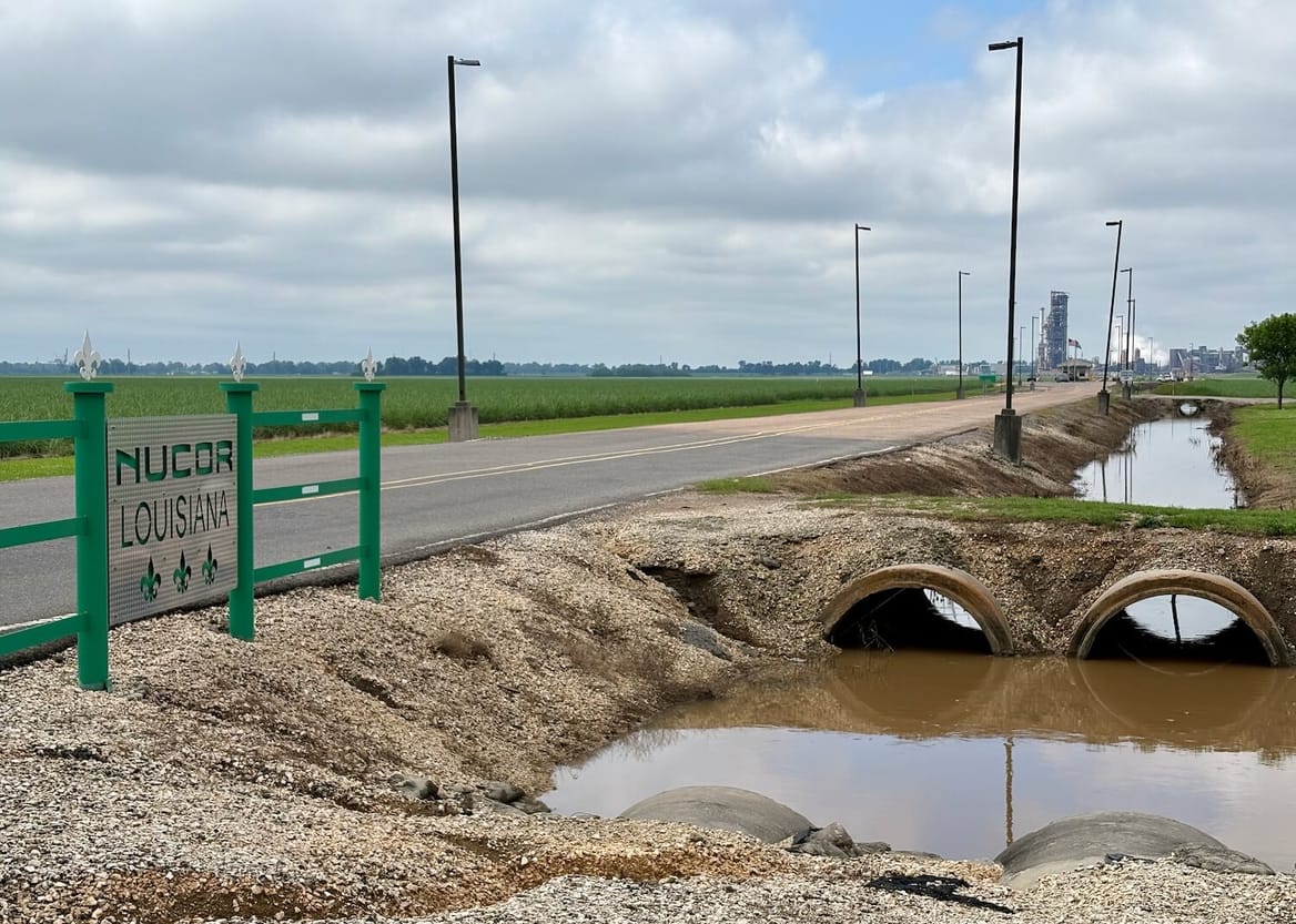 Green fence with "Nucor Louisiana" sign along a road and a ditch with water