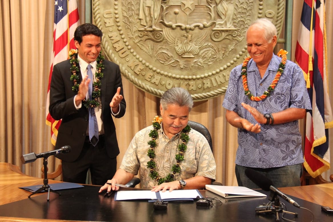 Three men, two standing and clapping; man in center with pen and document; state seal and two flags behind them