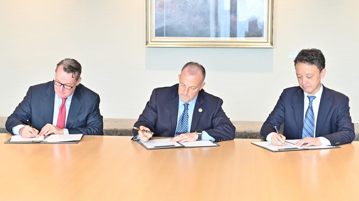 Three men in suits seated at a light-wood table, heads down signing documents