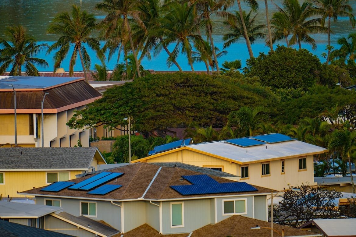 Homes of one and two stories with rooftop solar panels near a vibrant blue sea and lush trees, palm and others