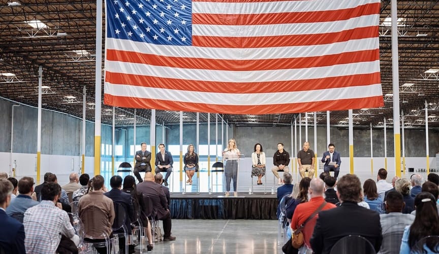Stage with speaker and others seated with a large US flag behind them and rows of people seated on a gleaming floor