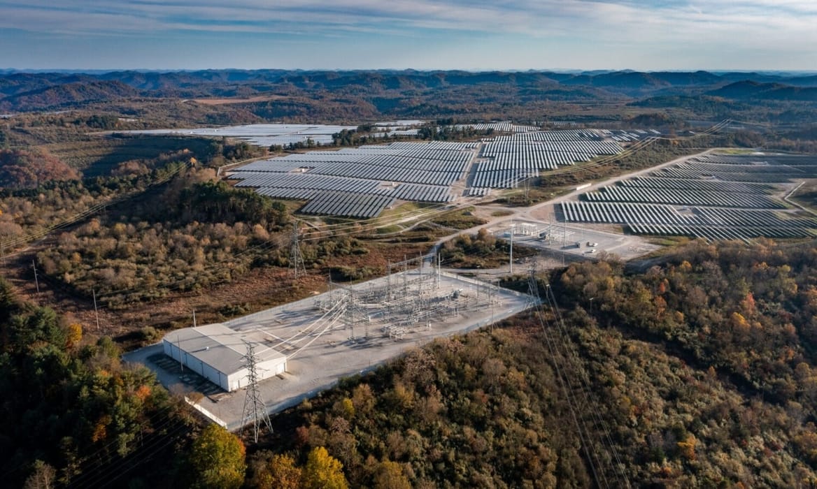 Overhead view of solar installations surrounded by forest and transmission equipment