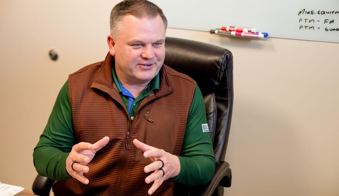 Man in rust-colored vest and green long-sleeved shirt in office chair with whiteboard behind