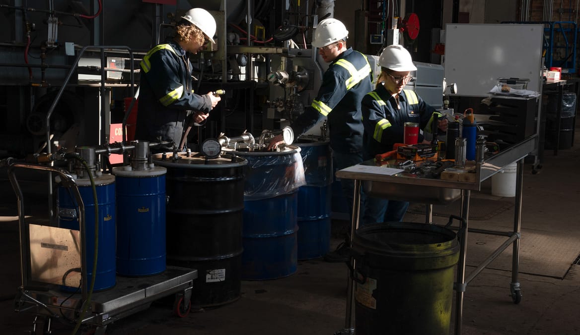 Three workers in dark lab coveralls and white hard hats