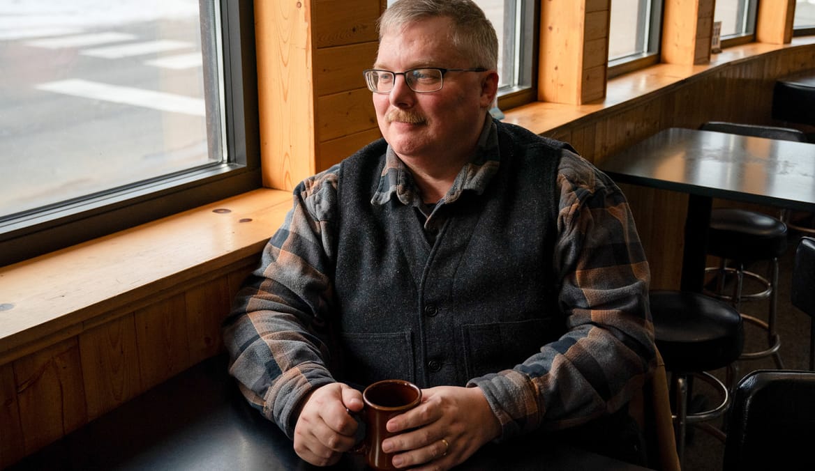 Man in plaid shirt and dark vest holding a mug while sitting at a restaurant table
