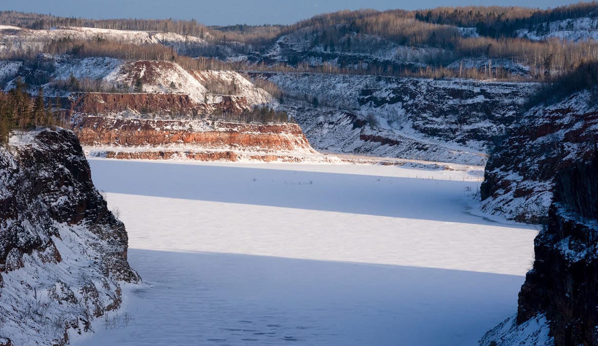 Open mine pit with steep sides dusted with snow and a snow or ice covered pond