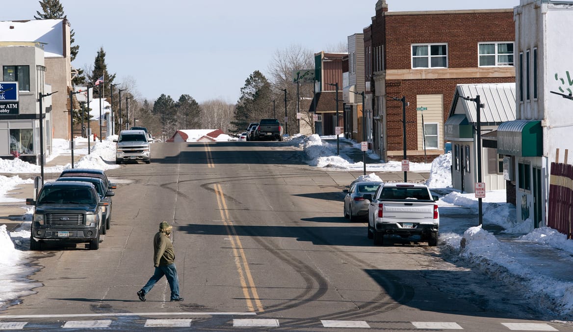 View of main street with one- and two-story buildings; on person crosing the street and a few parked cars