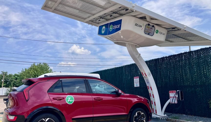 Red hatchback Zipcar parked and plugged in under a solar canopy held up with a curved white "arm"