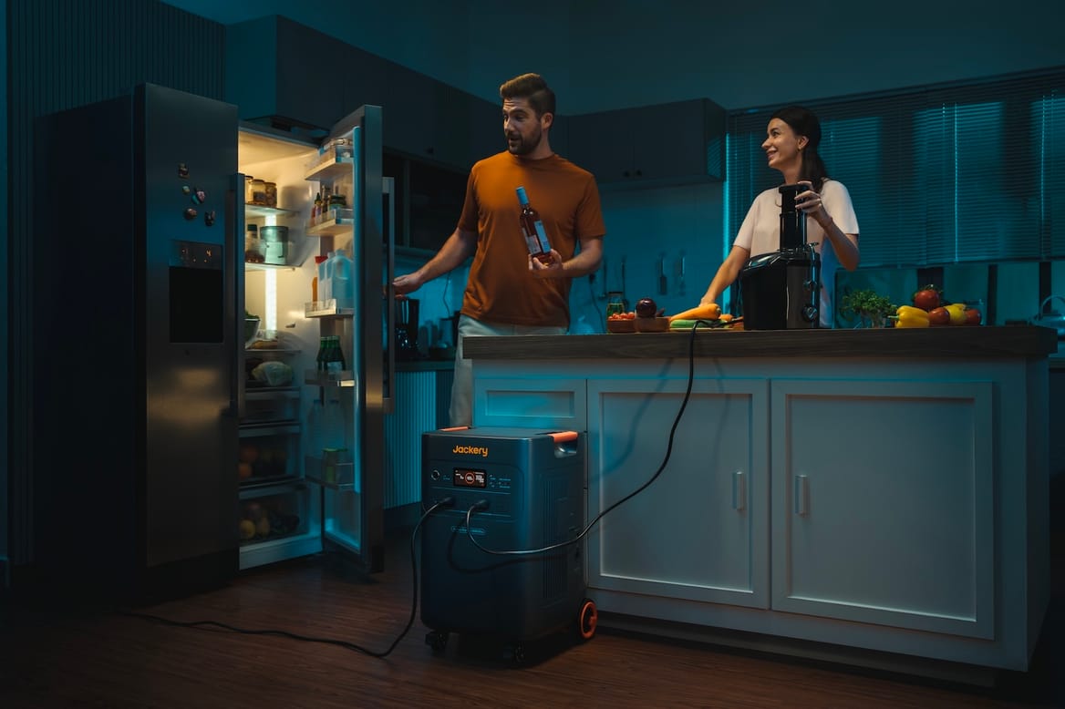 Two people in the kitchen use the fridge and a juicer — both plugged into a black portable battery — during a blackout