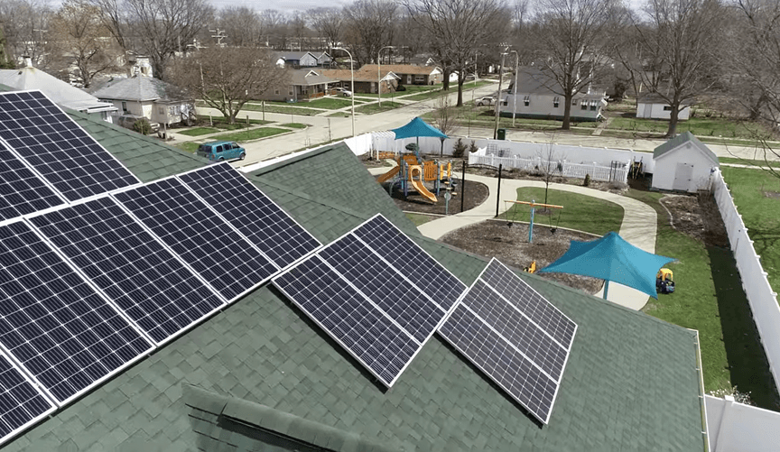Solar panels seen on a roof from above with playground equipment surrounded by a white fence on a lot