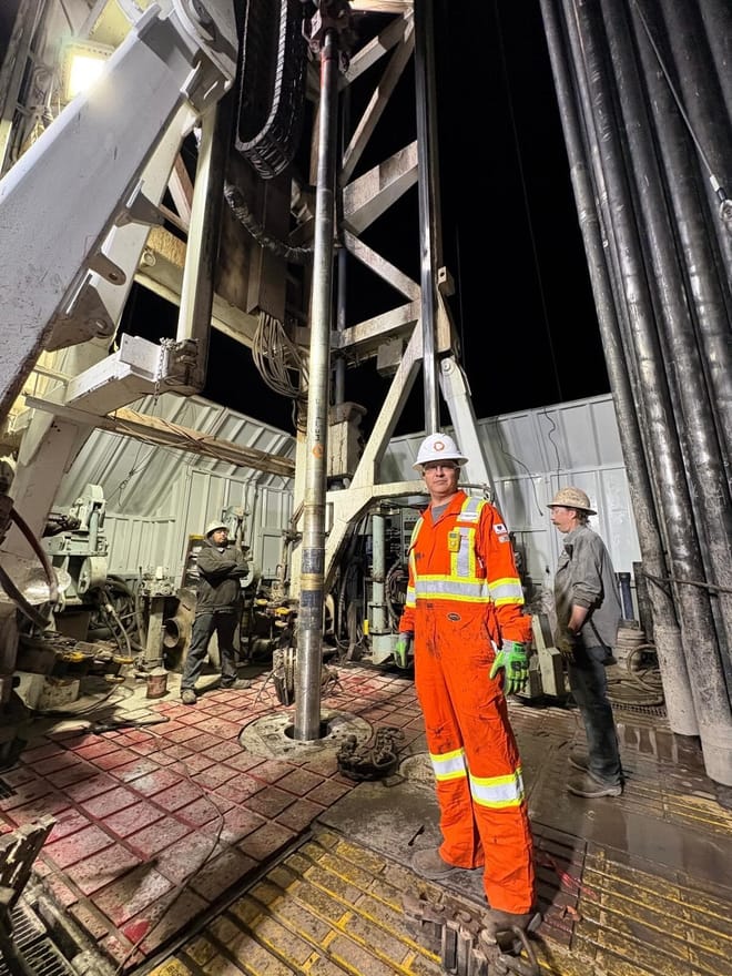 Man in orange worksuit and white hard hat by drilling equipmebnt