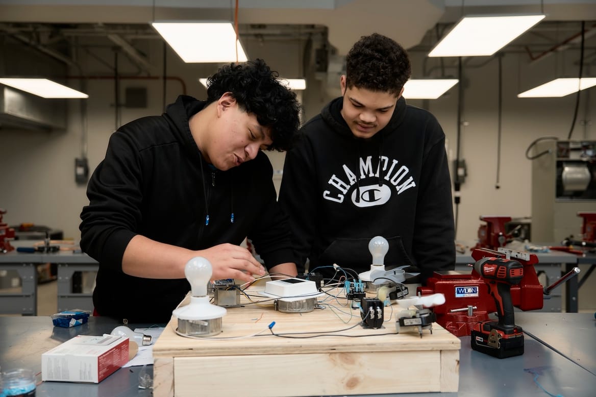 Two teens in black sweatshirts working with lightbulbs under fluorescent lights in a classroom