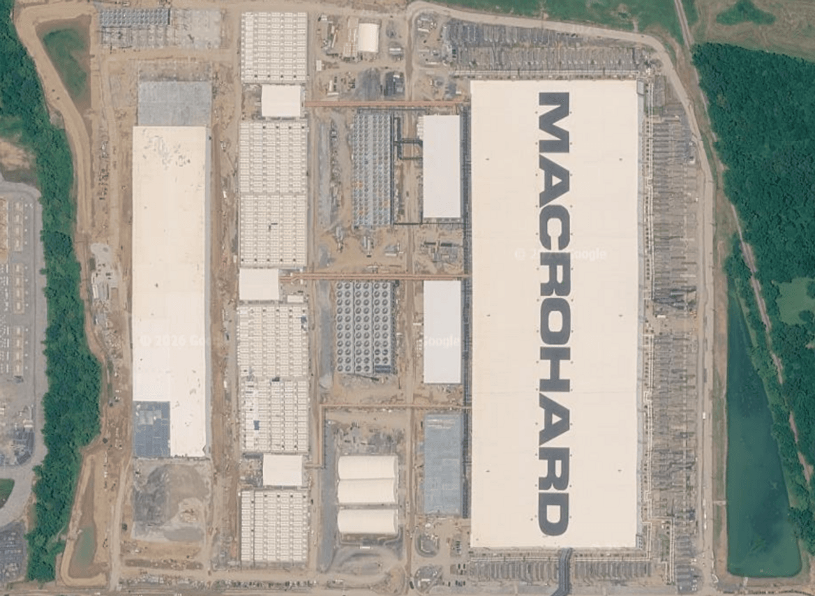 Aerial view of building with "Macrohard" in gray on the white roof flanked by other buildings rows of batteries, and parkiing