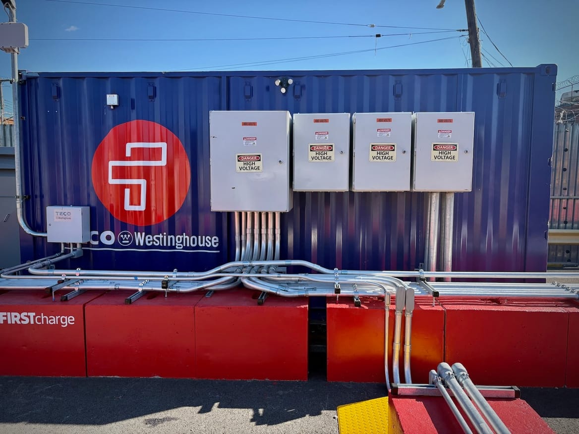 Blue battery (with "Westinghouse") on red base (with "First Charge"); metal pipes and boxes with "Danger: High Voltage" sings