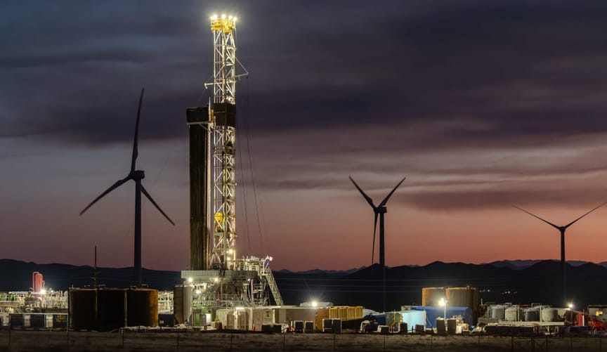 Geothermal plant with lights on at dusk or dawn with pink/orange sky and three wind turbines in background