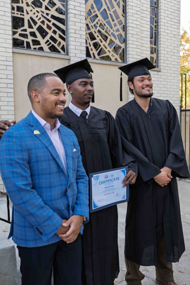A.J. Patton in blue jacket stands next to two graduates in black robes and hats with diploma