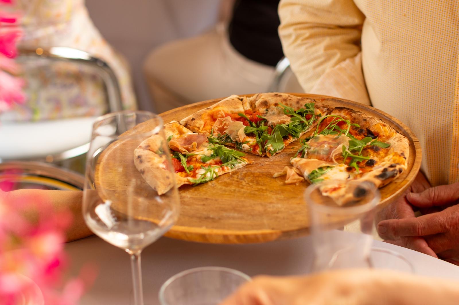 Partially eaten pizza with greens and ham on a wooden tray held by a person in a yellow shirt.