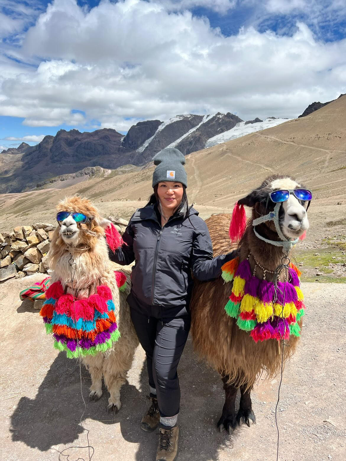 Travel nurse standing between two llamas decorated with colourful pom-poms and sunglasses, with mountains behind them