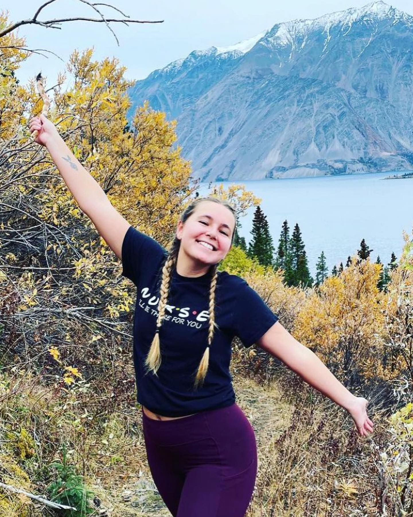 Travel nurse smiling with arms outstretched in front of a mountain lake and autumn trees
