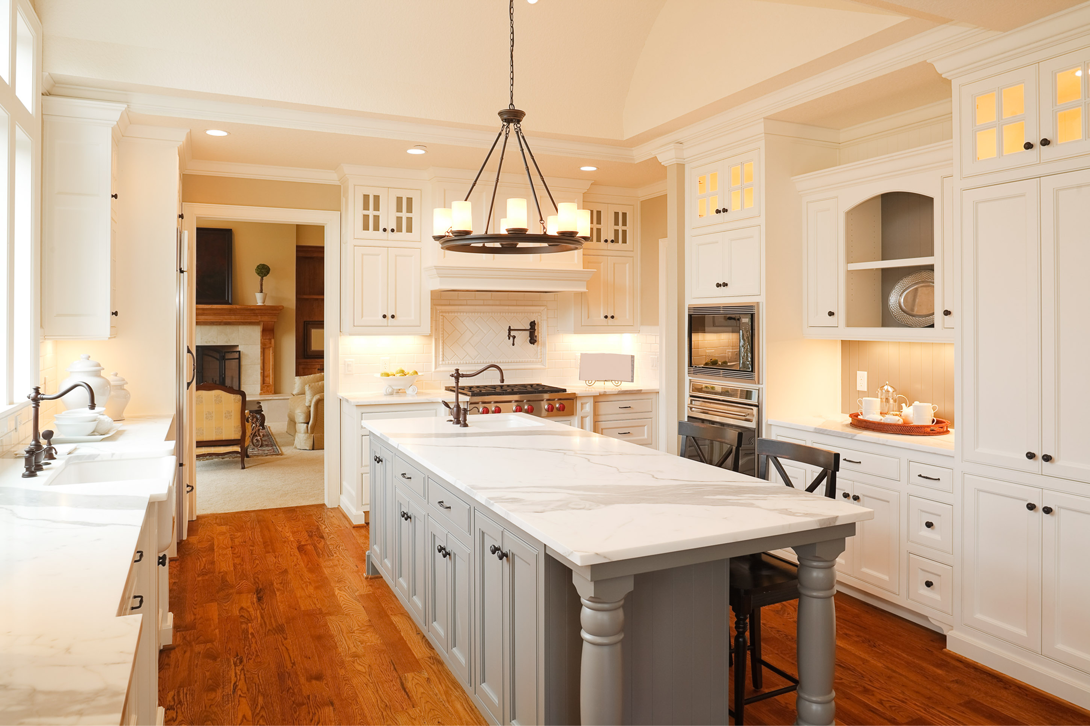 An elegant, traditional kitchen featuring creamy white cabinetry and a contrasting light grey island with decorative turned legs. The island and perimeter counters are topped with white marble. A large, dark metal circular chandelier with candle-style lights hangs in the center of the room. The space flows into a living room with a fireplace, and the floor is a rich, warm-toned hardwood.