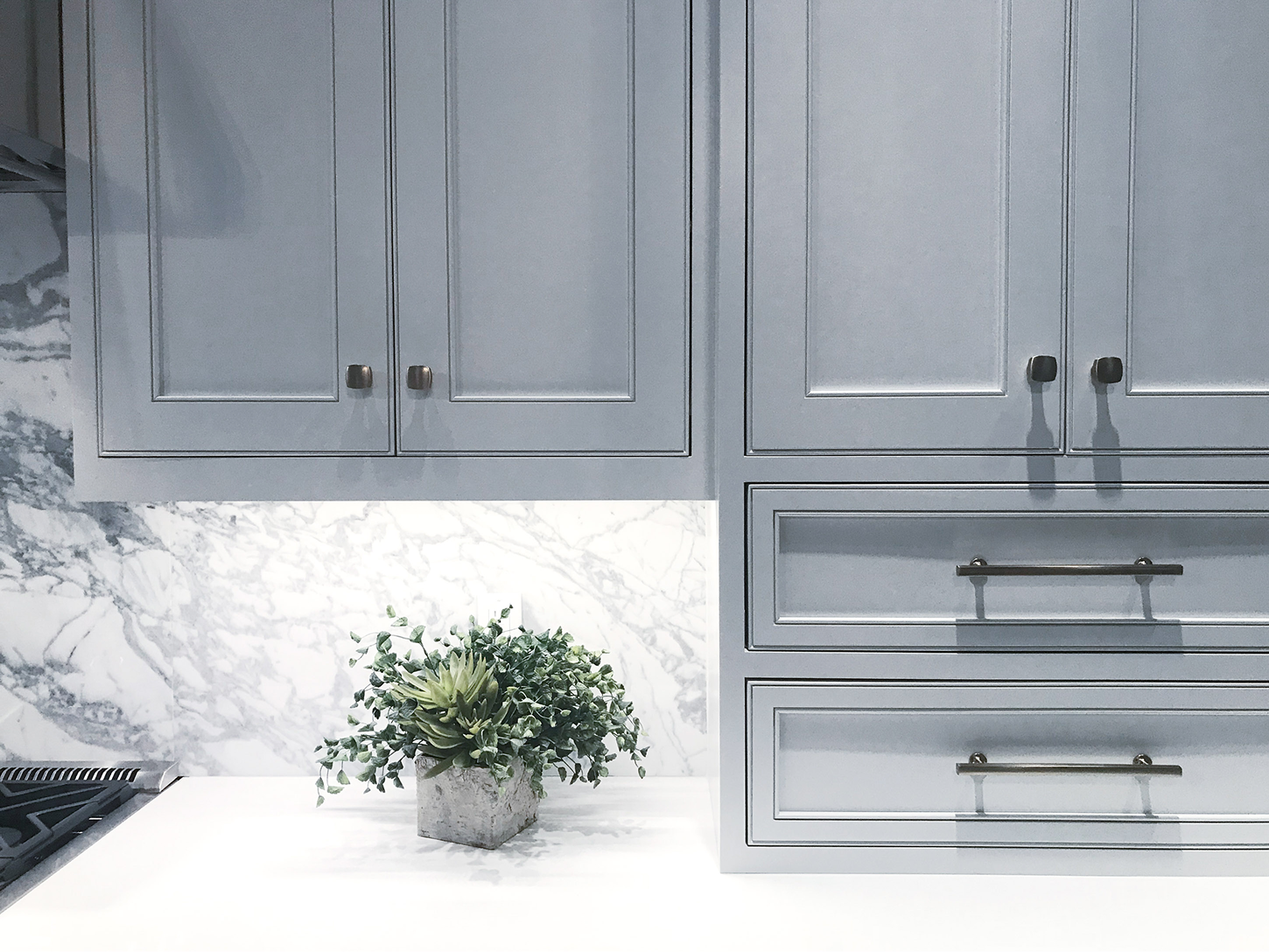 A close-up shot of light blue-grey Shaker-style kitchen cabinets with dark bronze hardware. The scene shows a white marble backsplash with heavy grey veining and a matching white countertop. A small green succulent in a grey stone pot sits on the counter, illuminated by bright under-cabinet lighting.