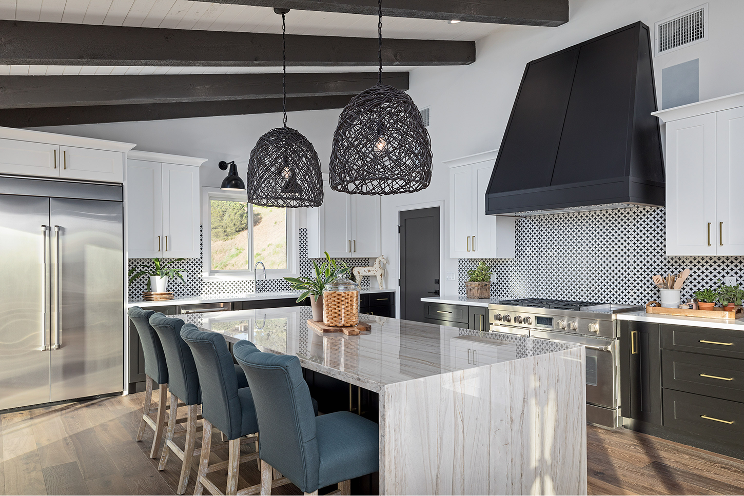 A contemporary kitchen with a high, vaulted ceiling and dark exposed wood beams. The design features a high-contrast palette with white upper cabinets, dark lower cabinets, and a bold black-and-white patterned tile backsplash. Two large, black woven basket pendant lights hang over a marble-topped island, where four teal upholstered chairs are tucked. A large black range hood sits above a professional stainless steel stove.