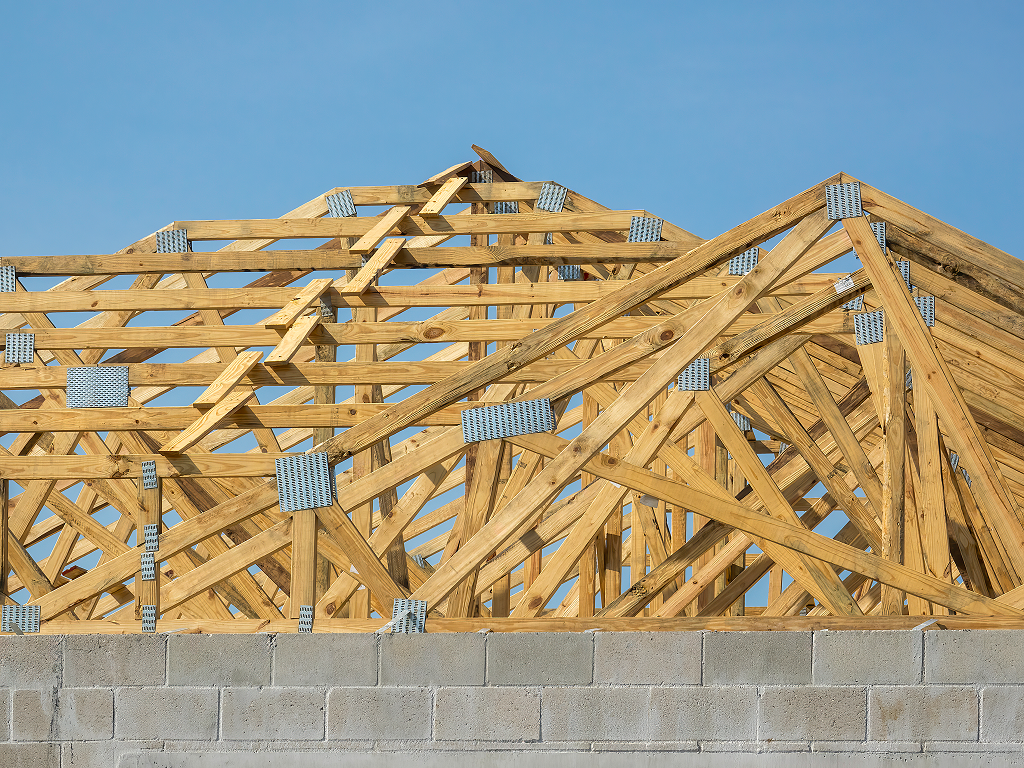 Wooden roof trusses and framing on a new construction house atop a concrete block wall under a clear blue sky.
