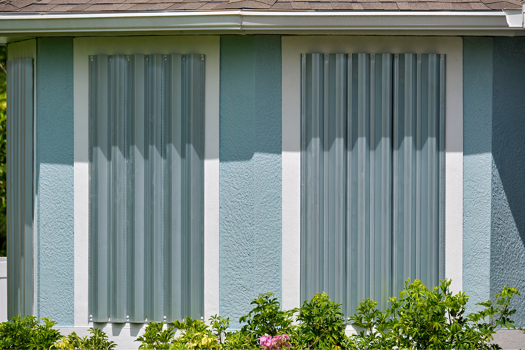 Corrugated metal hurricane shutters bolted over the windows of a light blue house with white trim and green shrubs.