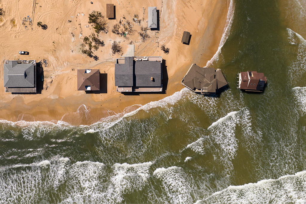 An aerial view of several coastal houses being overtaken by ocean waves and beach erosion.