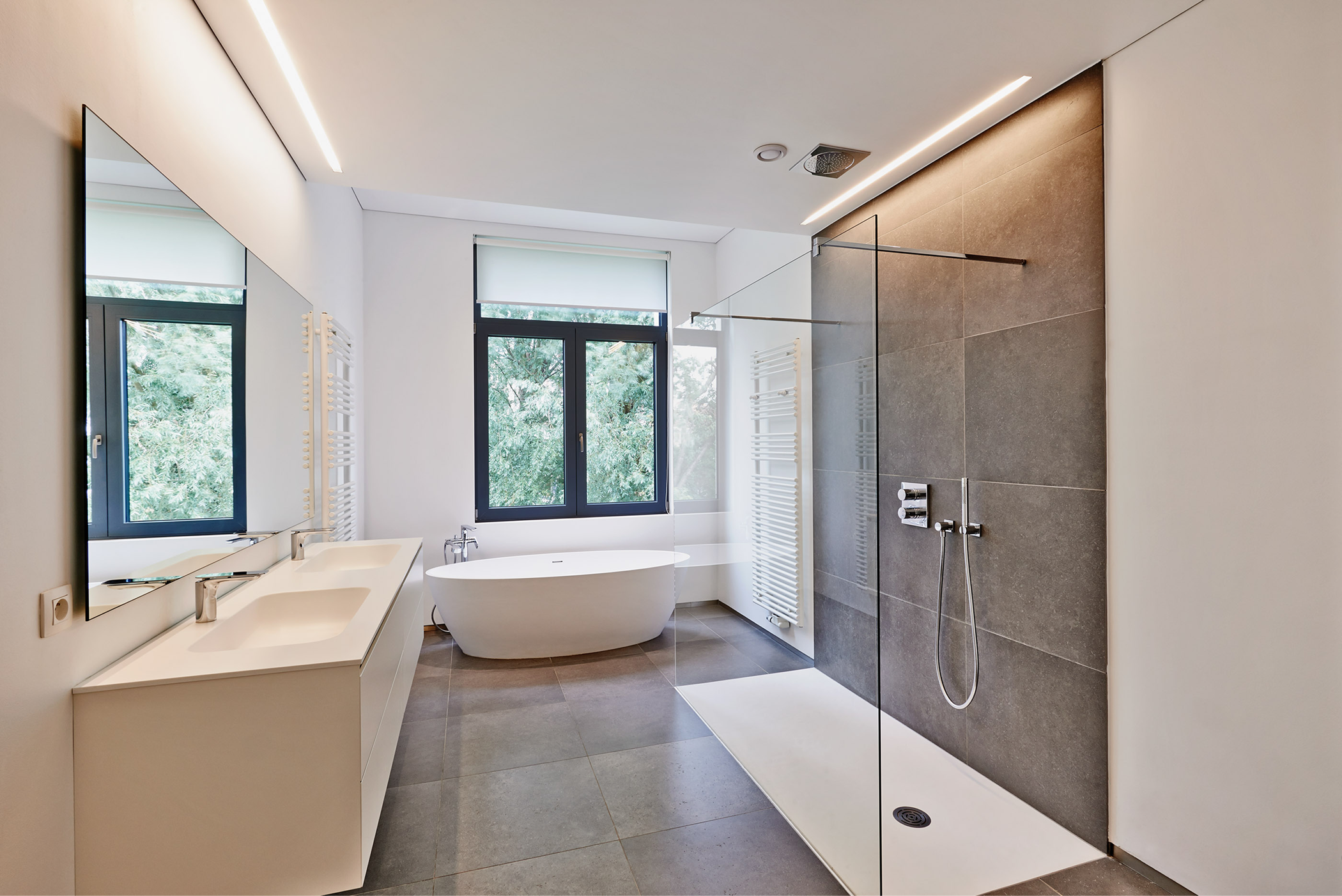 A minimalist luxury bathroom with a double vanity, a freestanding soaking tub by a window, and a large walk-in shower with dark grey wall tiles.
