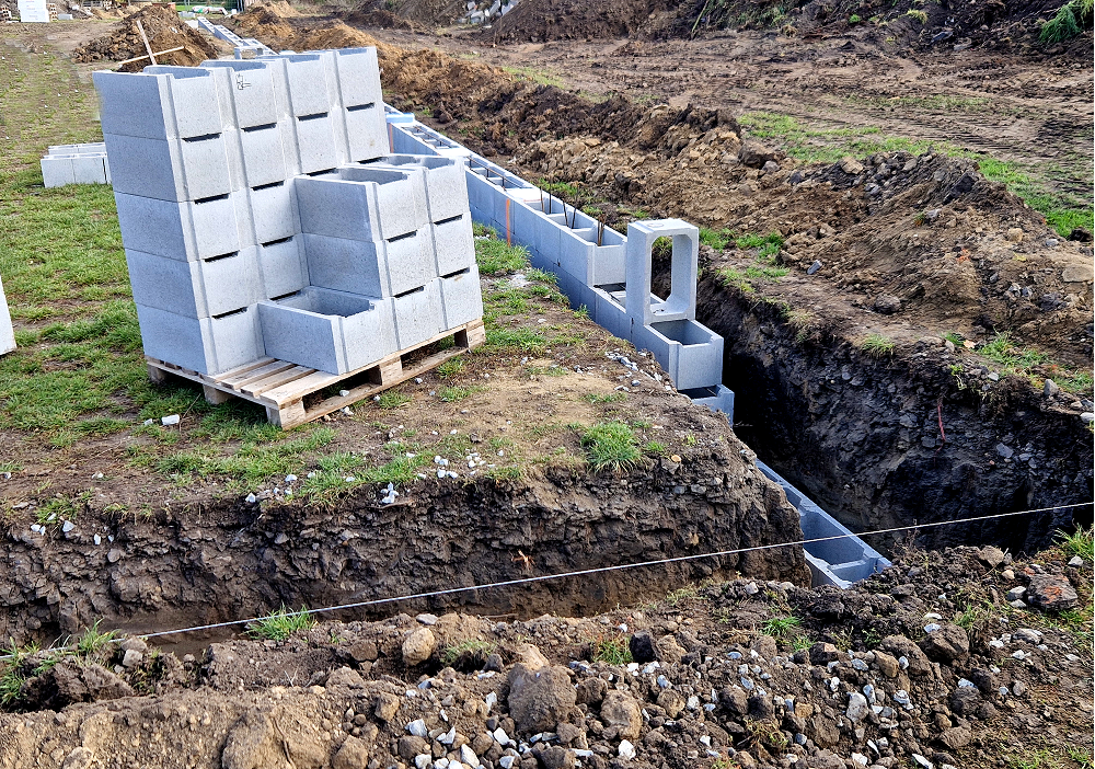 A construction site showing a deep dirt trench where concrete blocks are being laid for a building foundation, with a wooden pallet of extra blocks nearby.