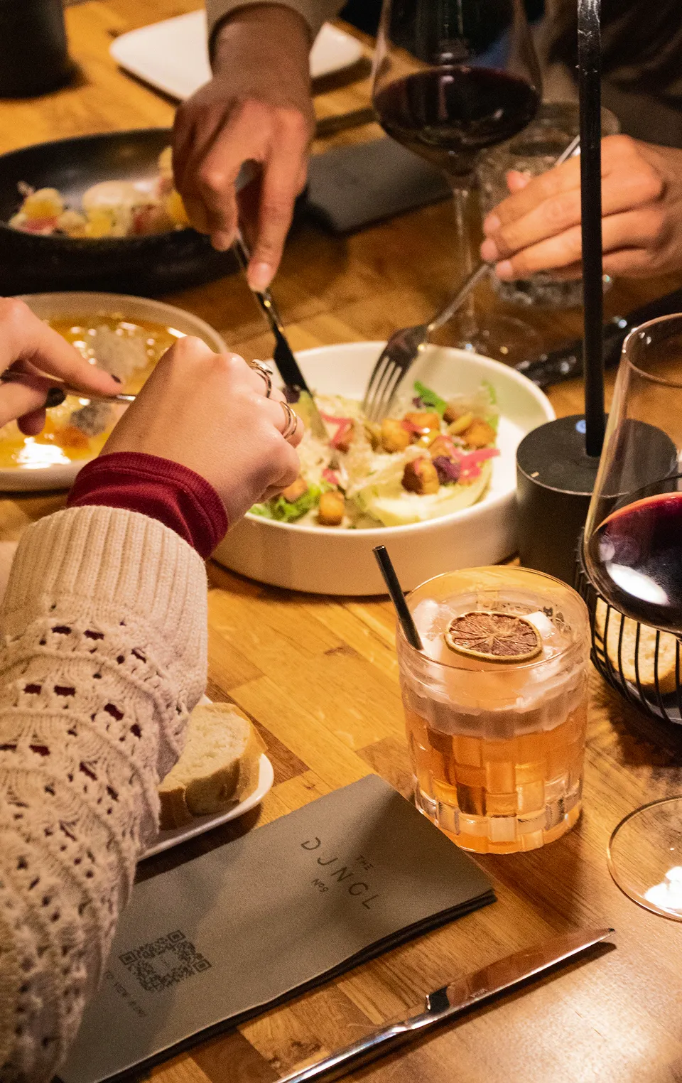 People dining together using utensils to eat food from plates with drinks on a wooden table.