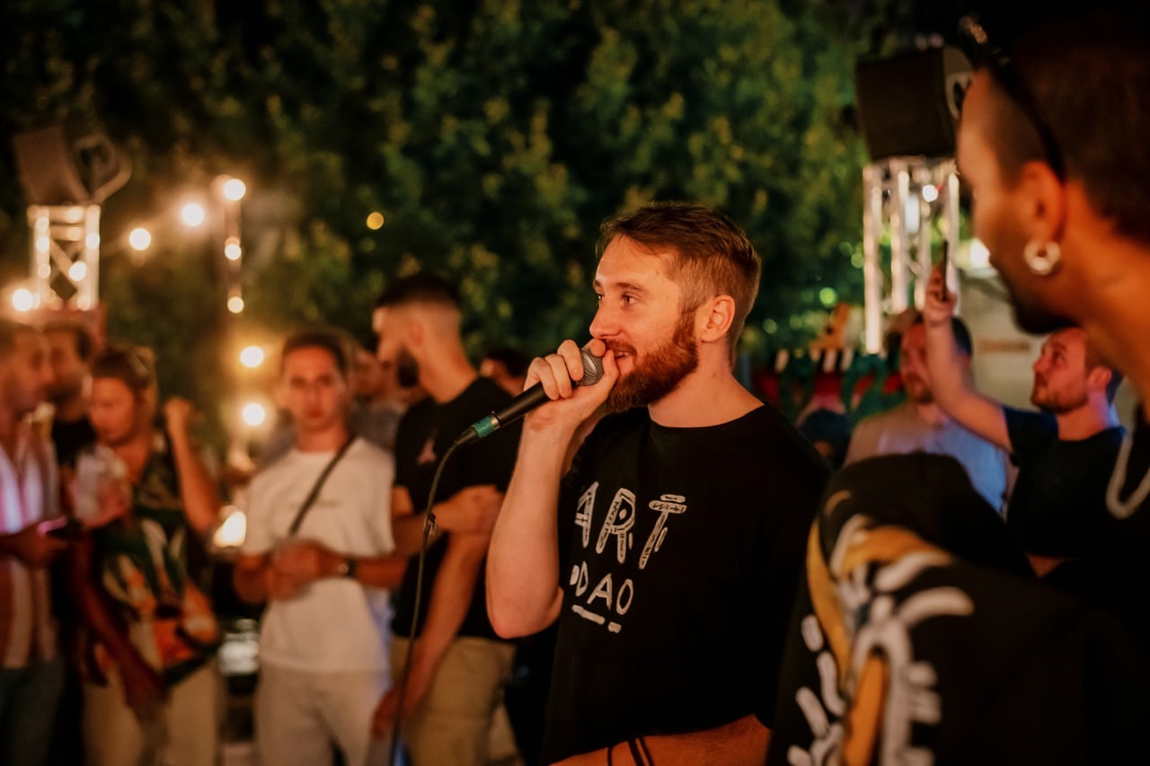 People standing around a man holding a mic. They are outside and appear to be having a great experience