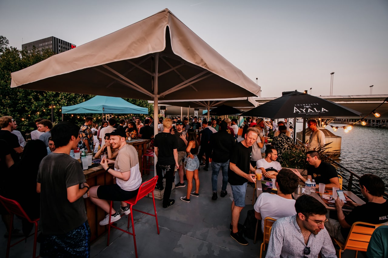a crowd of people on a patio next to the water socializing 