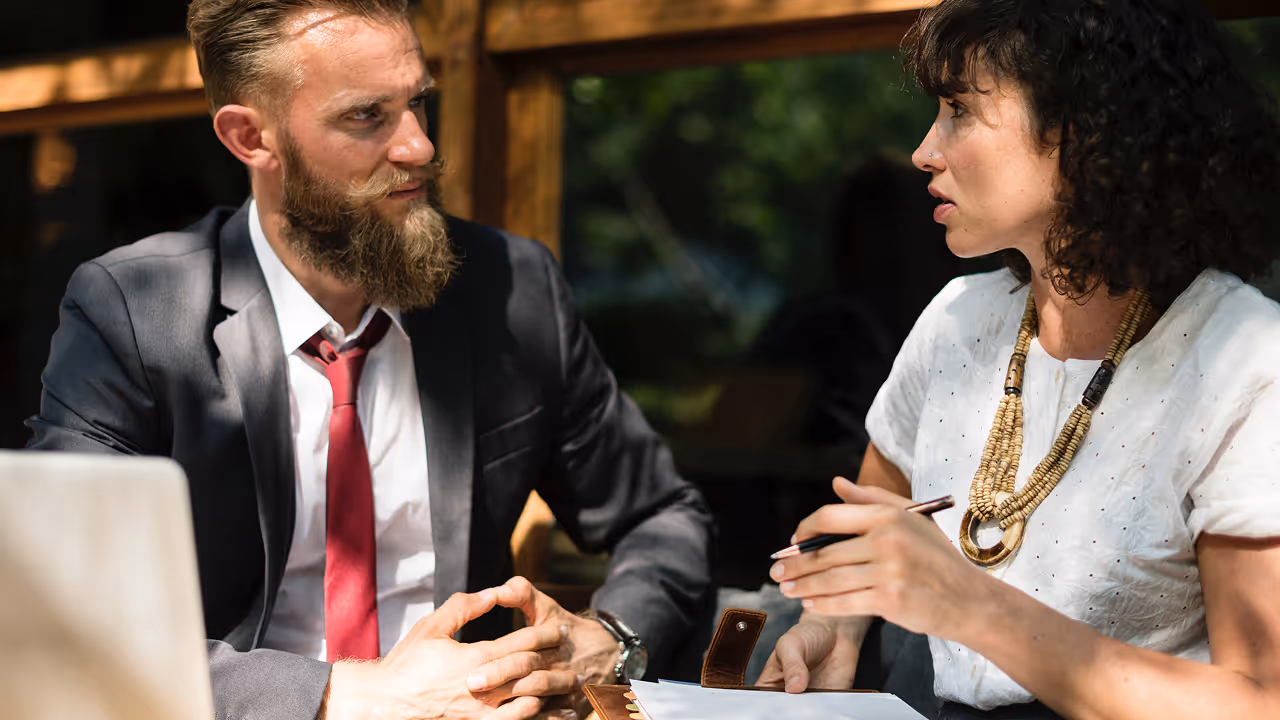 A man with a beard and a red tie is talking to a woman.