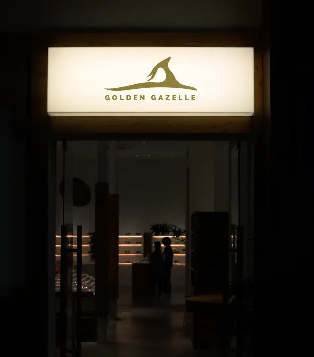 This image shows the illuminated storefront of **Golden Gazelle**, featuring its signature gazelle emblem and wordmark in muted olive green. The softly glowing signage contrasts beautifully against the dark exterior, drawing focus to the brand’s elegant simplicity. Inside, the warm ambient lighting and clean, minimal interior design create a refined and inviting atmosphere, reflecting Golden Gazelle’s identity—where modern craftsmanship meets timeless sophistication.
