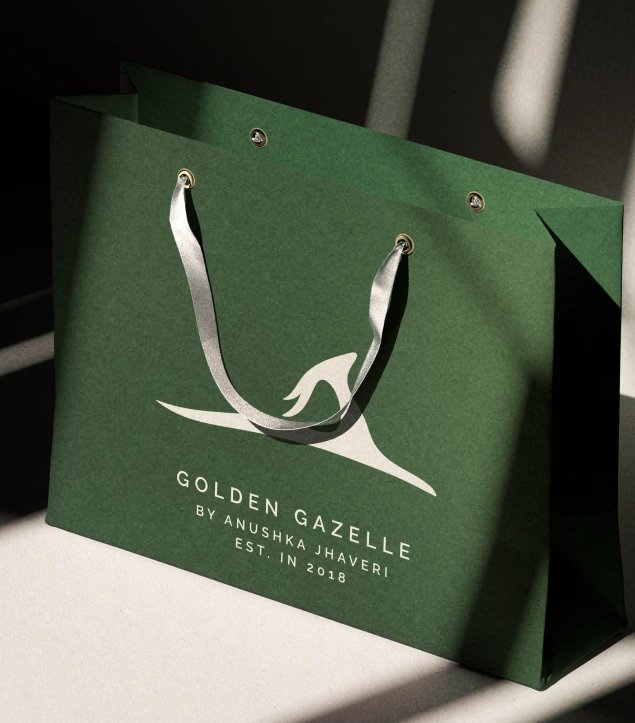 This image features a luxurious dark green shopping bag from Golden Gazelle by Anushka Jhaveri, established in 2018. The elegant matte finish of the bag is complemented by the brand’s signature gazelle logo in white, paired with satin silver handles that add a touch of refinement. The play of light and shadow enhances the texture, reflecting Golden Gazelle’s ethos of timeless elegance, craftsmanship, and understated luxury. The design exudes sophistication and trust, perfectly embodying the brand’s premium jewellery experience.
