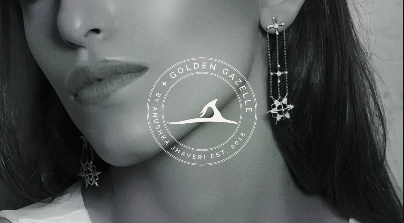 This monochrome photograph features a woman wearing elegant statement earrings from Golden Gazelle by Anushka Jhaveri. The image captures a close-up of her lower face, lips, and earrings with soft lighting and a refined grayscale treatment, highlighting the brand’s signature craftsmanship and minimal, sophisticated aesthetic. The Golden Gazelle logo sits at the center, blending seamlessly into the composition, reinforcing the brand’s identity of modern luxury rooted in timeless design.
