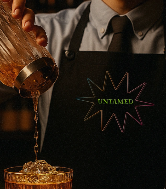 Bartender in a black apron with a colorful starburst logo reading ‘UNTAMED’ pours a golden cocktail into a glass over ice, representing the artistry, precision, and bold spirit of Untamed Gin’s mixology-driven brand identity.