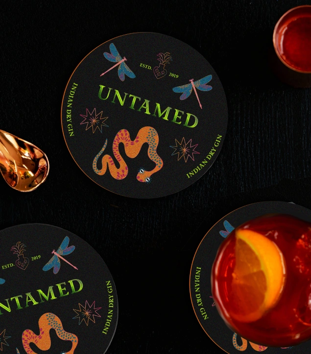 Branded coasters for Untamed Gin featuring mystical illustrations of snakes, dragonflies, and celestial motifs in vivid colours on a dark background, reflecting the brand’s bold, exotic, and craft-inspired identity for premium Indian dry gin
