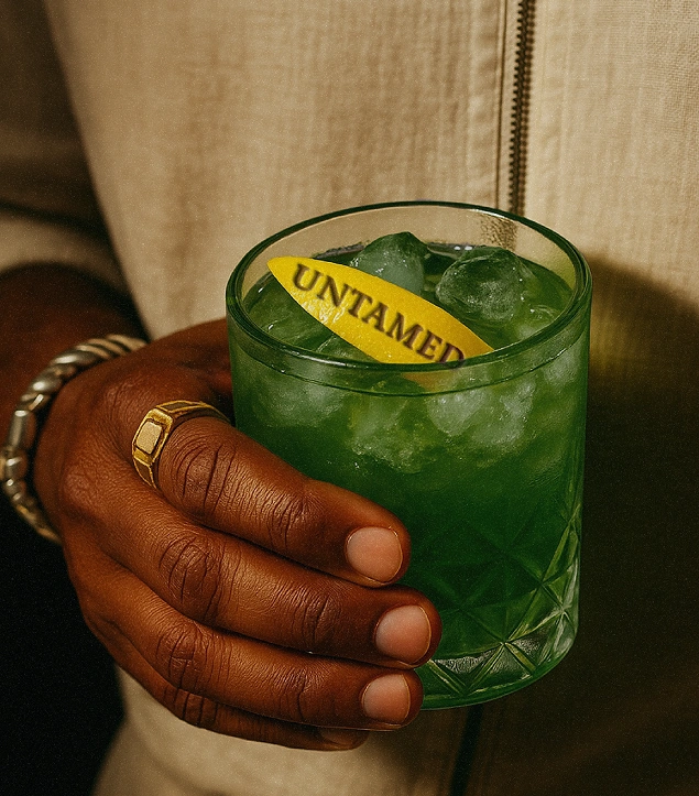 Beverage photography for Untamed Gin by Dot and Grid – close-up of a hand holding a green craft cocktail with a branded lemon peel garnish reading ‘Untamed,’ showcasing premium alcohol branding and lifestyle imagery for a luxury gin label