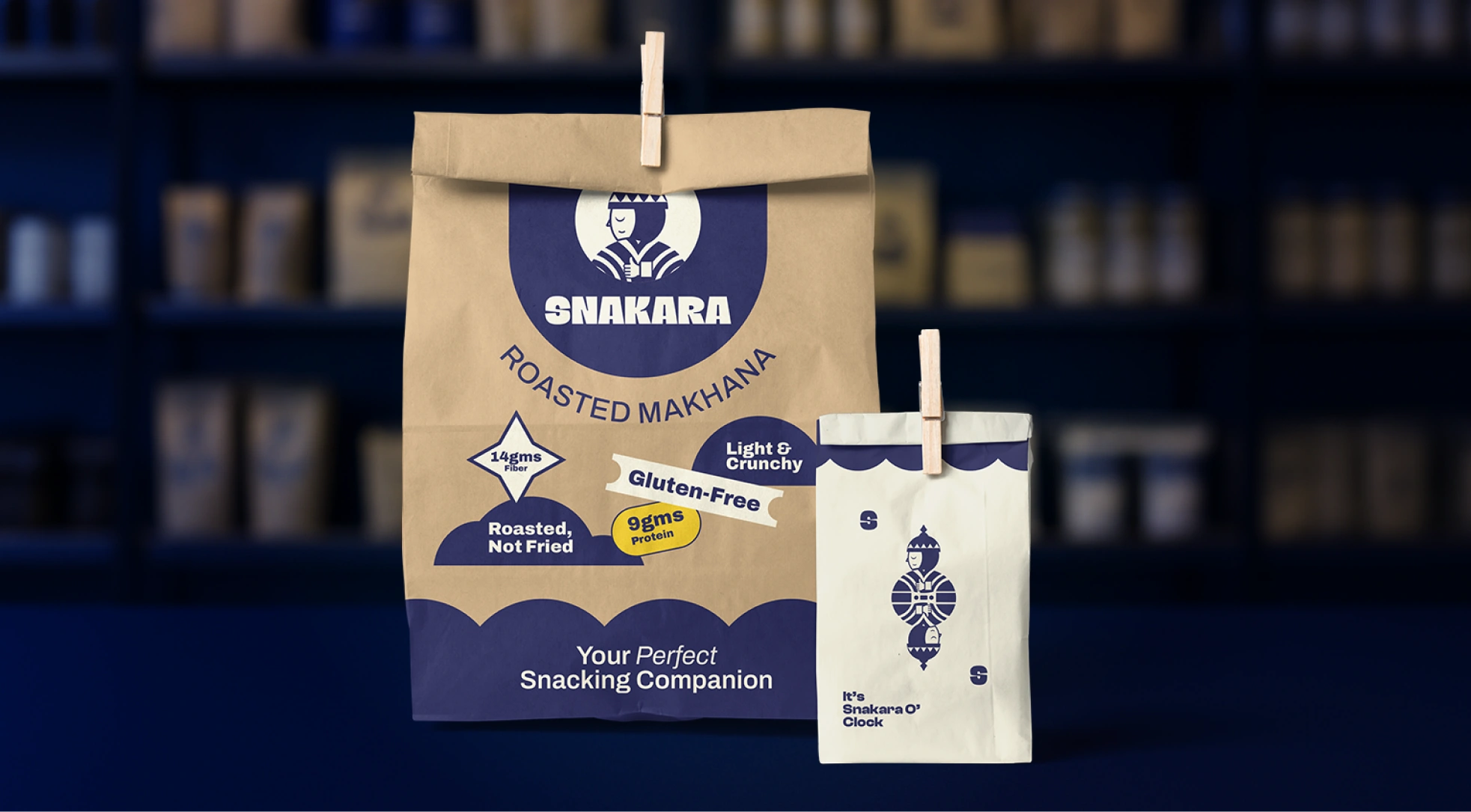 This image features **Snakara’s eco-friendly paper packaging**, showcasing the brand’s earthy yet modern design approach. The two-toned kraft and cream color palette blends natural authenticity with bold minimalism. The larger bag highlights Snakara’s roasted makhana range with playful graphic badges—*Gluten-Free, Light & Crunchy, 14g Fiber, 9g Protein, Roasted Not Fried*—while the smaller pouch carries the brand’s signature Samurai mascot with the phrase *“It’s Snakara O’Clock.”*

Together, they express Snakara’s philosophy of mindful snacking with joy and purpose. The packaging feels both artisanal and fresh, positioning the brand as an everyday healthy indulgence wrapped in thoughtful, sustainable design.
